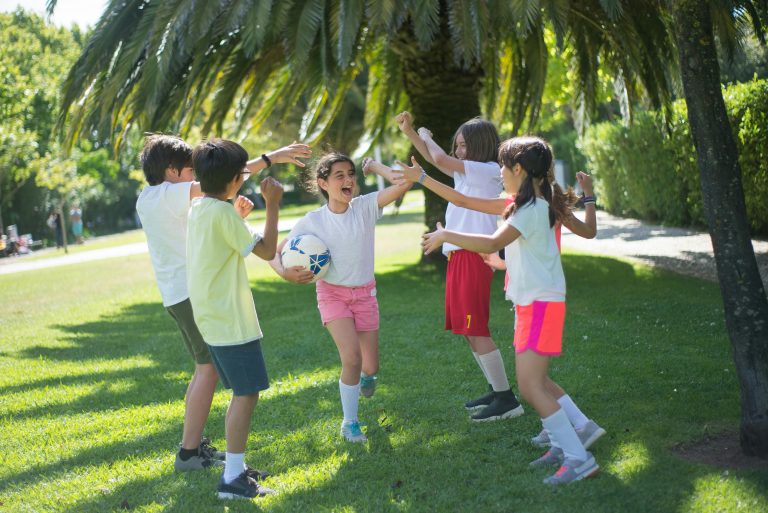 Group of happy children playing football under a palm tree in a sunny park.